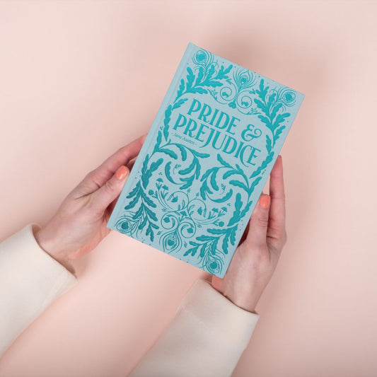 Hand holding a blue book titled 'Pride & Prejudice' against a light pink background. This is the illustrated wordsworth edition.