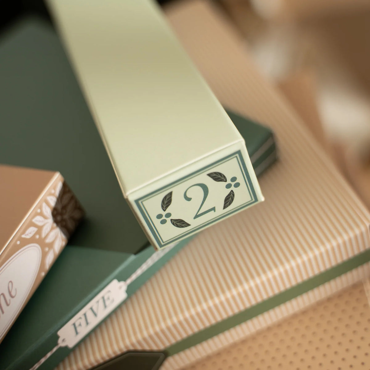 A close-up of a long rectangular box with a pale sage green finish and a decorative label showing the number 2, placed on top of other gift boxes in a softly blurred setting.