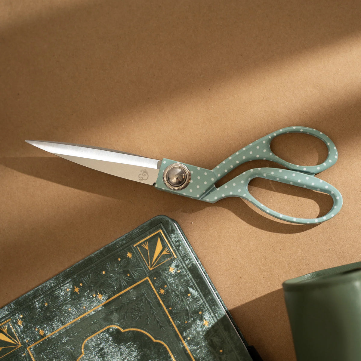 A pair of scissors with sage green handles featuring white polka dots and silver stainless steel blades, resting on a brown textured surface next to a decorative green notebook.
